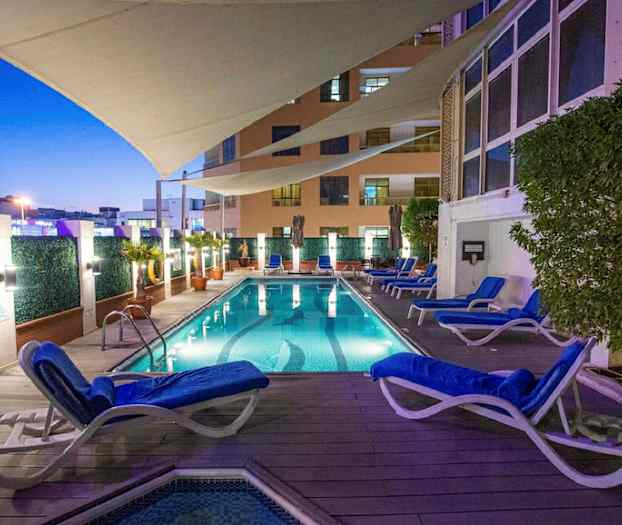 En upplyst swimmingpool med omkringliggande solstolar och växter, under ett skyddande segel på kvällen. (AI-genererad text)