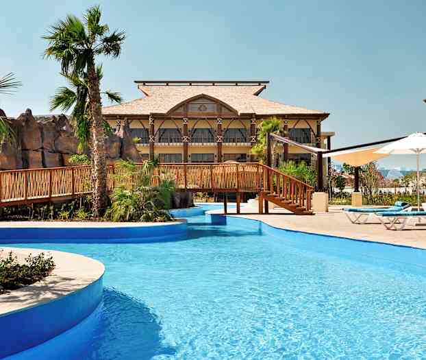 En pool med blått vatten och en träbro, omgiven av palmer och byggnader. (AI-genererad text)