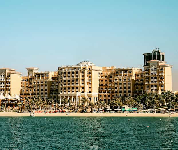En stor hotellbyggnad vid en strand med palmer framför, en ljus himmel i bakgrunden och havet i förgrunden. (AI-genererad text)