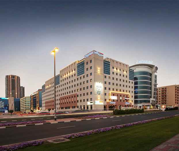 En stadsmiljö vid skymning med flerbostadshus och moderna hotellbyggnader. En väg med biltrafik och blommor längs trottoaren. (AI-genererad text)