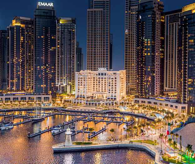 En stadssilhuett vid kvällen med upplysta höga byggnader och en småbåtshamn i förgrunden. (AI-genererad text)