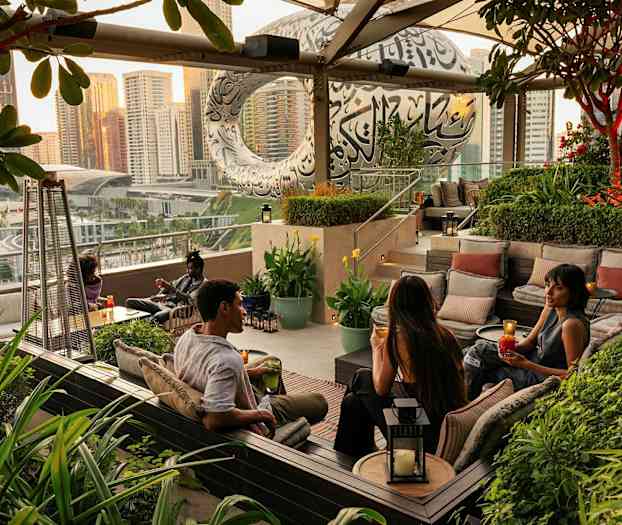 En utomhuslounge med sittgrupper omgiven av grönska, med en stadsvy och en modern byggnad i bakgrunden. (AI-genererad text)