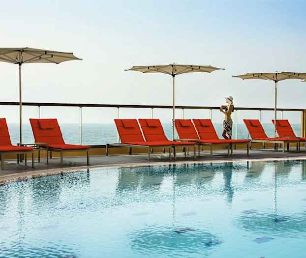 En utomhuspool med röda solstolar och vita parasoller vid sidan av, med havsutsikt i bakgrunden. (AI-genererad text)