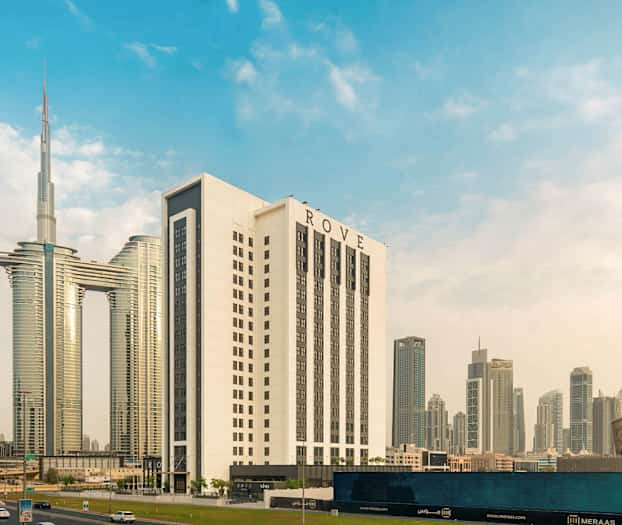 Ett modernt stadsmotiv med flera höga byggnader, däribland ett större vitt hotell med namnet ROVE, omgiven av andra skyskrapor och arkitektoniska strukturer. (AI-genererad text)