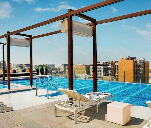 En utomhuspool med utsikt över en stad. Solstolar, bord och dekorationer är placerade runt poolen. (AI-genererad text)