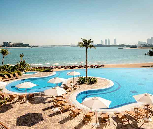 En hotellpool med solstolar och parasoller belägen vid en strand med utsikt över havet och en stad i bakgrunden. (AI-genererad text)