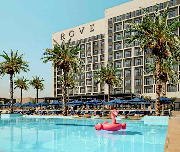En byggnad märkt 'ROVE' bakom en pool med palmer och solstolar runtom samt en rosa flamingo uppblåsbar leksak i vattnet. (AI-genererad text)
