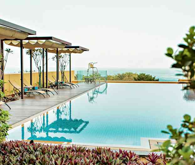 En avbildning av en utomhuspool med solstolar under skuggande tak, omgiven av växtlighet och hav i bakgrunden. (AI-genererad text)