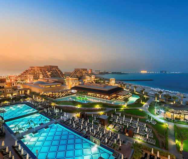 Vy över ett hotellområde med byggnader, pooler och strand vid havet. (AI-genererad text)