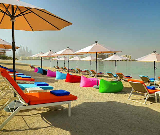 En sandstrand med solstolar utrustade med färgglada dynor och parasoller, vid en lugn vattenyta. (AI-genererad text)