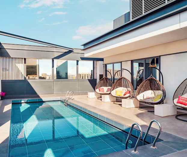 En takterass med en liten pool, hängande flätade stolar och moderna omgivningar. Byggnader syns i bakgrunden. (AI-genererad text)