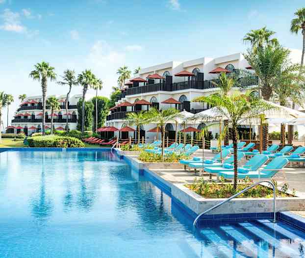 Ett hotellområde med flera våningar, palmer, en swimmingpool och solstolar under parasoll. (AI-genererad text)