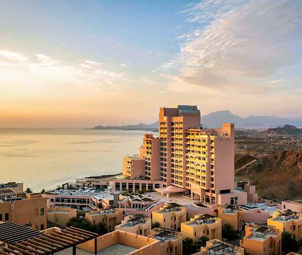 Hotellbyggnad nära havet vid solnedgången, omgiven av mindre byggnader och berg i bakgrunden. (AI-genererad text)