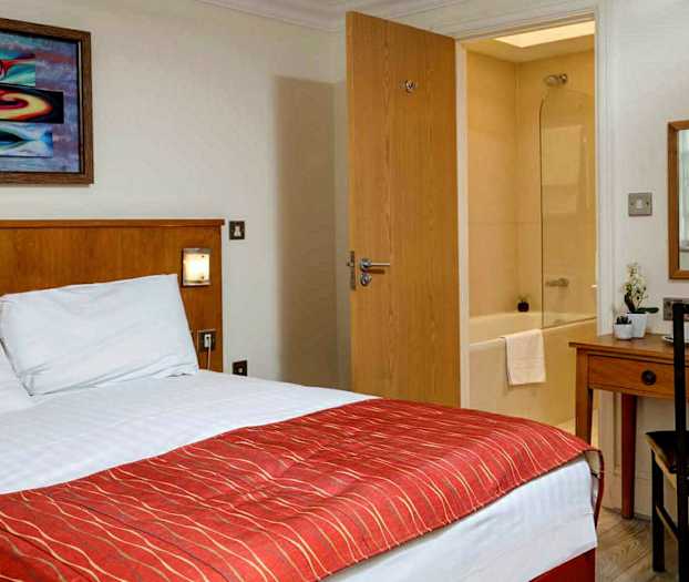 Hotellrum med säng, skrivbord, spegel och vy mot ett badrum. (AI-genererad text)