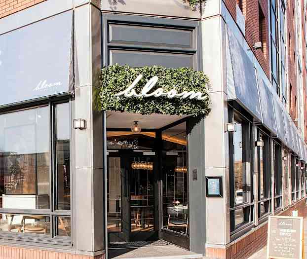 Entrén till restaurangen Bloom med stora fönster och en skylt med namnet omgiven av grönska. (AI-genererad text)