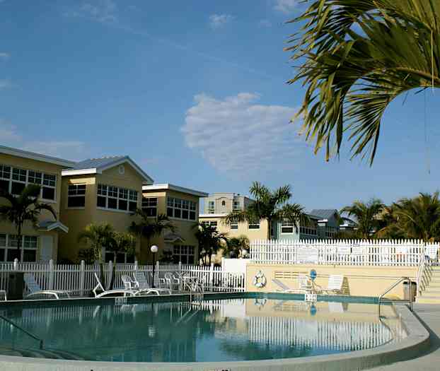 En pool omgiven av solstolar, parasoller och hus, med tropiska palmer i bakgrunden. (AI-genererad text)