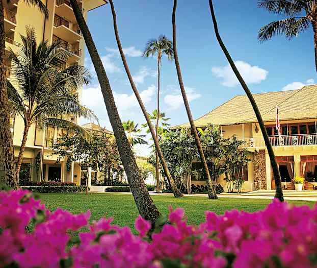 En byggnad omgiven av grönska med palmer och blommor i förgrunden. (AI-genererad text)