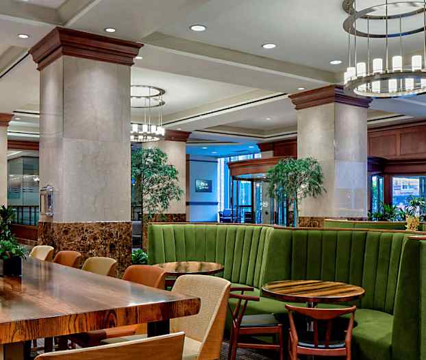 Ett modernt möblerat interiör med gröna bås-sittplatser och träbord, omgiven av växter och belysning. (AI-genererad text)