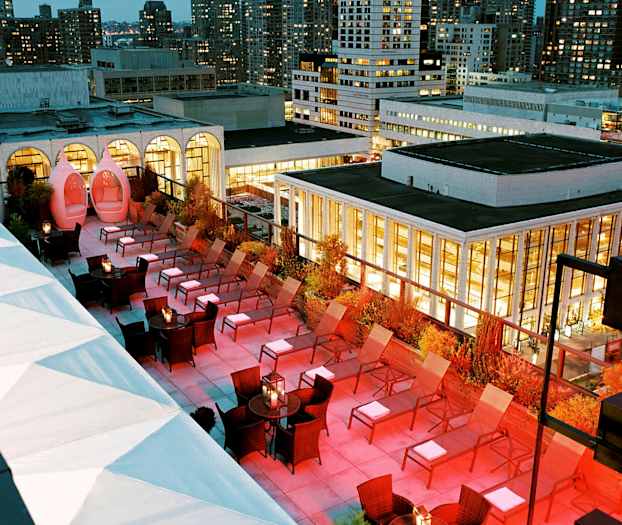 En takterrass med solsängar och belysning under kvällen, omgiven av byggnader. (AI-genererad text)