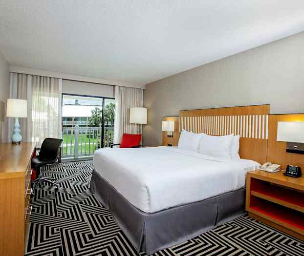 Ett hotellrum med en stor säng, arbetsbord och stol, samt en balkong med utsikt över en gräsmatta. (AI-genererad text)