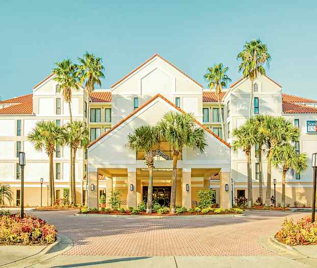 Bild av ett hotell med en vit fasad, röda takpannor, omgivna av palmer och välskötta trädgårdar. (AI-genererad text)