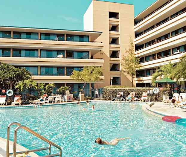 En stor swimmingpool omgiven av solstolar och en fler vånings byggnad i bakgrunden. (AI-genererad text)