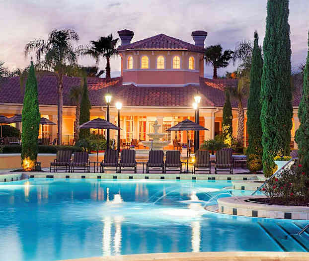 En utomhuspool med omgivande solstolar och parasoller framför en upplyst villa i skymningen. (AI-genererad text)