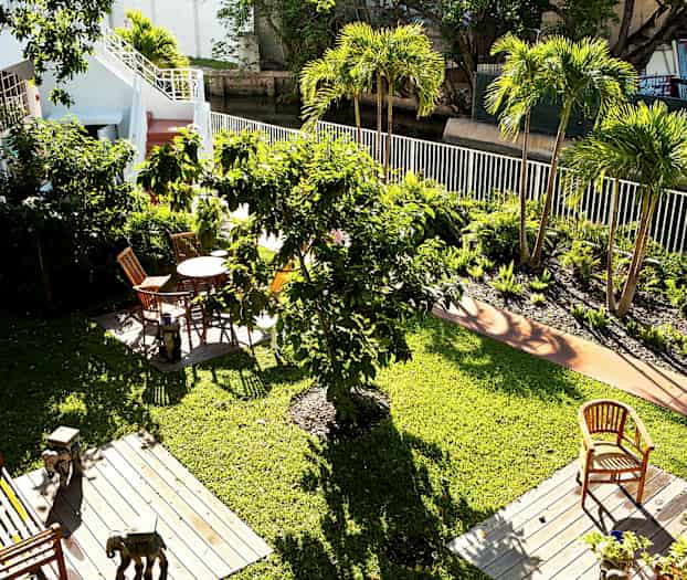 Vy över en grön trädgård med flera träd och blomsterrabatter. Det finns trädgårdsmöbler på träplattformar placerade på gräsmattan. (AI-genererad text)