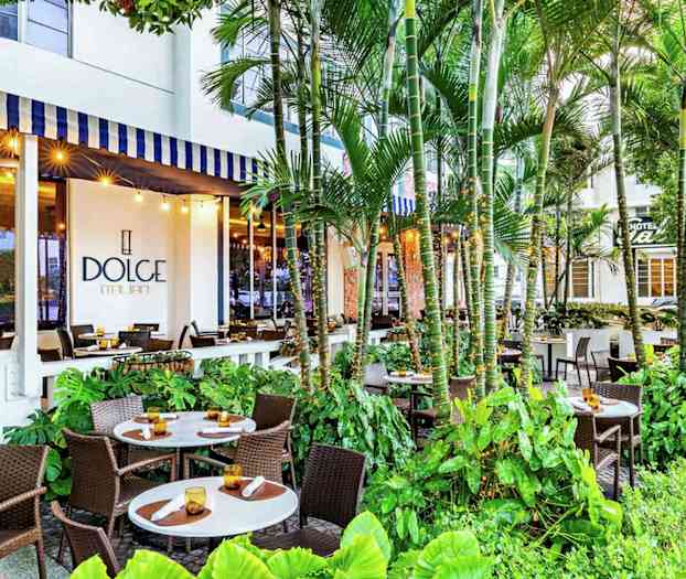 En utomhusrestaurang med flera bord och stolar omgivna av frodig grönska, med en skylt för 'Dolce Italian' synlig på byggnaden. (AI-genererad text)