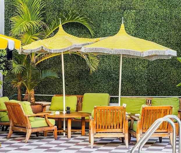 En utomhusstol med gröna sittdynor och träbord under gul- och vitrandiga parasoller, med växtlighet i bakgrunden. (AI-genererad text)