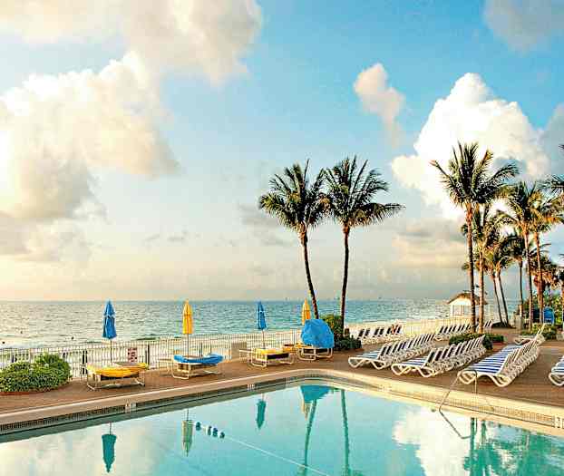 En pool med omgivande solstolar och parasoller vid havet, med palmer i bakgrunden. (AI-genererad text)