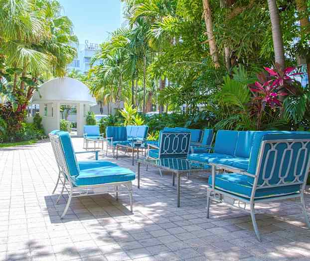 Utemöbler med blå dynor står uppställda på en stenlagd uteplats, omgiven av palmer och grön växtlighet. (AI-genererad text)