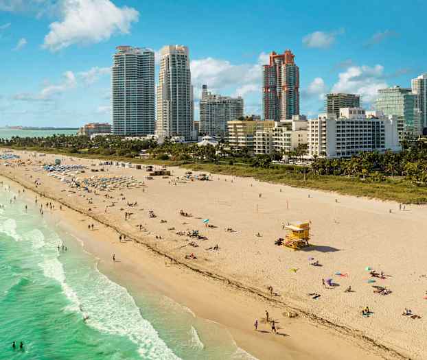 En sandstrand med några människor som badar och solar nära havet. Höga byggnader syns i bakgrunden. (AI-genererad text)