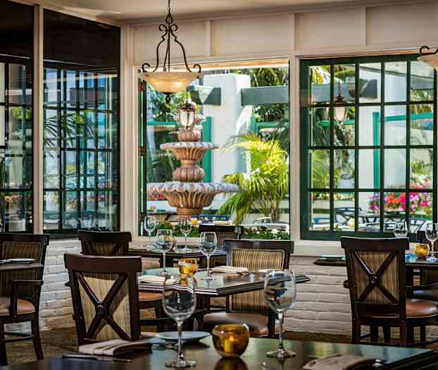 Bild på en restaurang med bord som står uppdukade, stora fönster släpper in ljus och visar en uteplats med en fontän och gröna växter. (AI-genererad text)
