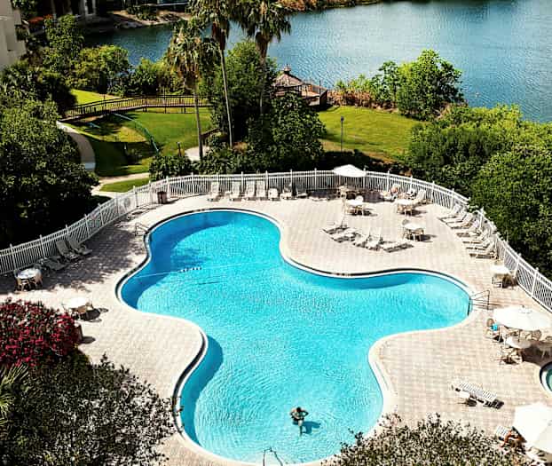 En utomhuspool omgiven av solstolar och bord med parasoller vid en sjö. (AI-genererad text)