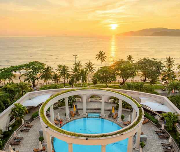 En vy av en pool omgiven av pelare och växter. I bakgrunden syns en strand med palmer och hav vid solnedgången. (AI-genererad text)