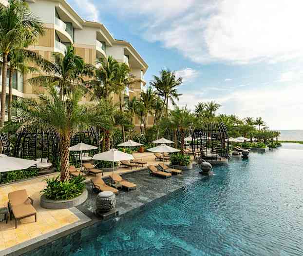 Hotellpool som omges av solstolar, parasoller och palmer med havsutsikt. (AI-genererad text)