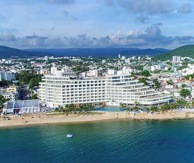 En modern byggnad vid en strand med havet i förgrunden och en stad i bakgrunden. (AI-genererad text)