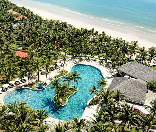 Bilden visar en översiktsvy av ett tropiskt semesterområde med en stor pool omgiven av palmer, solstolar, och parasoller nära en sandstrand vid havet. (AI-genererad text)
