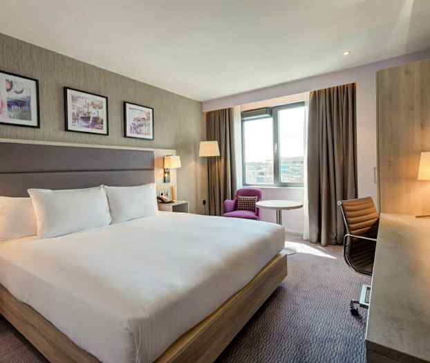 Ett hotellrum med en stor dubbelsäng, skrivbord, stol och en fåtölj vid fönstret. (AI-genererad text)