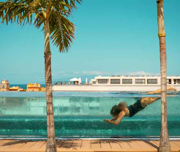 En person simmar under vattnet i en genomskinlig pool, med byggnader och en palm mot en blå himmel i bakgrunden. (AI-genererad text)