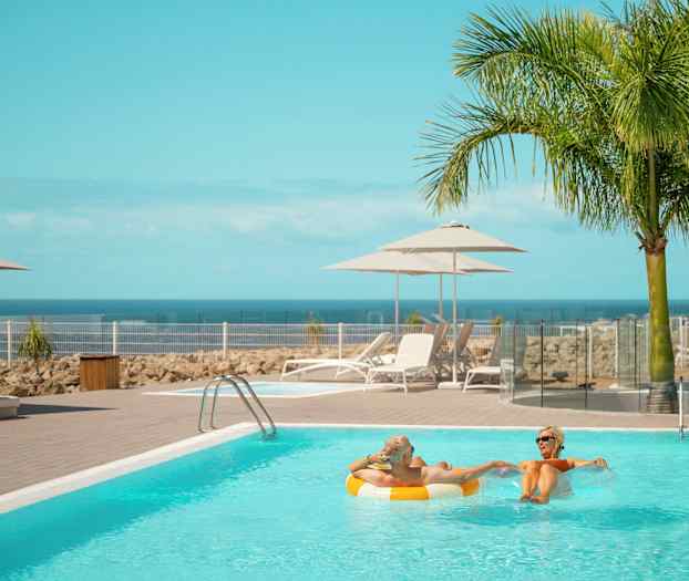 En pool med solstolar och parasoller bredvid, mot en bakgrund av hav och palmer. (AI-genererad text)