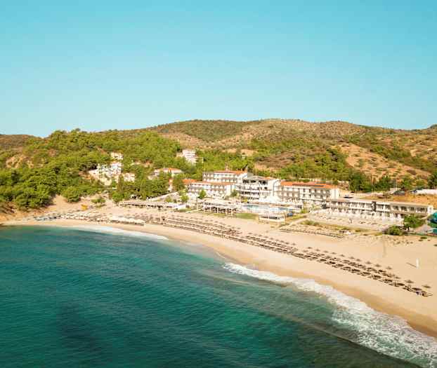 Flygfoto av en strand, hotellbyggnader och grönskande sluttningar. (AI-genererad text)