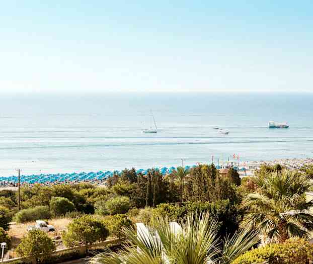 Utsikt över en strand med blå parasoller, omgiven av gröna träd. Flera båtar syns på det lugna havet. (AI-genererad text)
