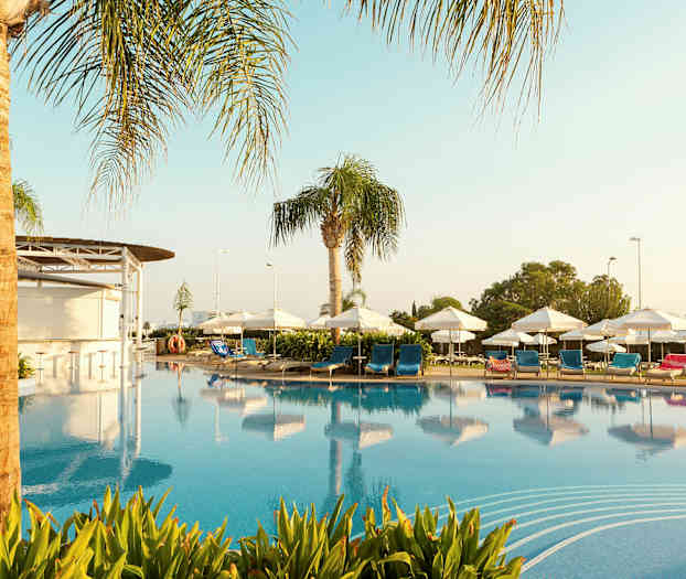 En pool omgiven av solstolar med blå och röda kuddar, parasoller och palmer. (AI-genererad text)