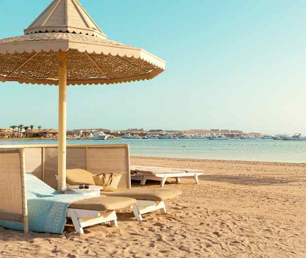 En strand med solsängar och parasoll, samt båtar på havet. (AI-genererad text)