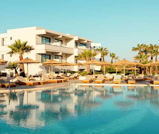 Hotell med vit fasad bakom en swimmingpool och solstolar med parasoller. (AI-genererad text)