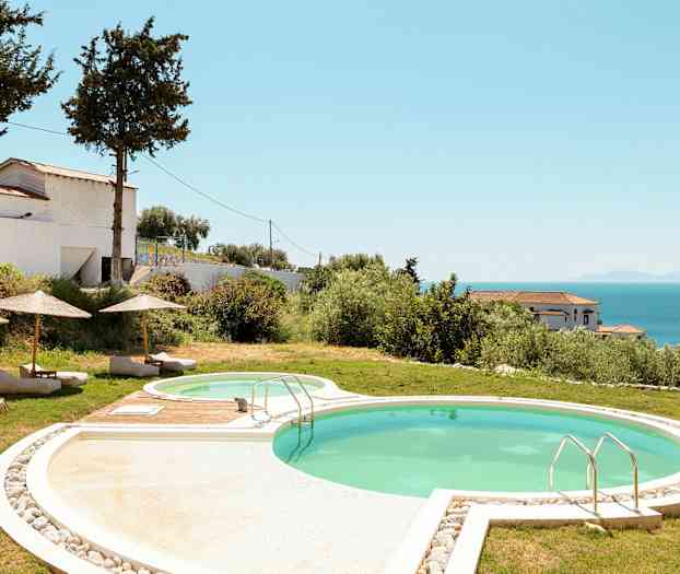 En utomhuspool med solstolar och parasoller, omgiven av gräs och växtlighet. Havet och byggnader syns i bakgrunden. (AI-genererad text)