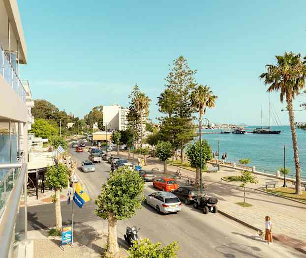 En stadsgata som går bredvid havet, med bilar, palmer och några byggnader i bakgrunden. (AI-genererad text)