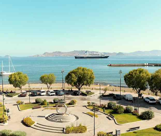 Vy över en hamn med parkeringsplats och träd i förgrunden, båtar och ett kryssningsfartyg på det blå havet, samt berg i bakgrunden. (AI-genererad text)